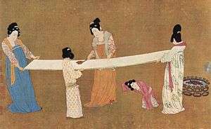 Two women hold out a long bolt of white silk by the corners, while two other women brush out the silk with combs.