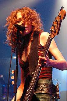Melissa Auf der Maur singing into a microphone while playing a bass guitar.