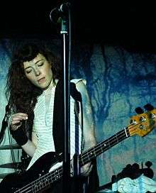A female musician performing with a bass guitar against a black and blue backdrop. A microphone is visible above her.