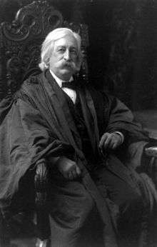 A man in his mid-fifties with bushy white hair, sideburns, and a mustache. He is sitting in a chair wearing a black judicial robe over a black suit and tie and white shirt.