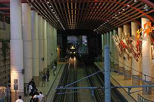A train enters an enclosed train station with two side platforms. The platforms are decorated with pillars and a piece of artwork.