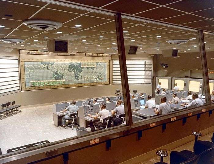 A large room seen through windows at the rear, with two rows of people seated at desks and computers, and a map taking up the whole of the front wall.