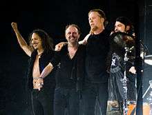 Four men on a stage with their arms behind one another; all are wearing dark clothing