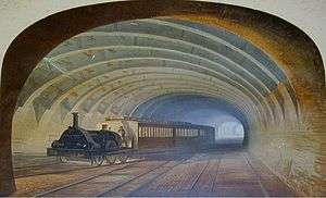 A well-lit railway tunnel recedes into the distance. A train with a steam locomotive and carriages is heading towards the viewer but taking the branch off to the left.