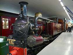 A steam tank locomotive is shown indoors, funnel towards the viewer, in purple livery. A large pipe connects the pistons at the front with the side tank