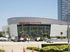 A white round building that has one glass front wall showing with a sign in red text that reads "Staples Center" in capital letters. In the background, there are multiple people waling in front of the building and a white parked car and a cloudy blue sky.