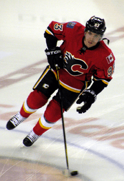 A hockey player in a red uniform with a black stylized "C"-logo on his chest makes a sharp turn while skating on the ice.