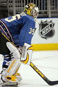 An ice hockey goaltender facing the right of the camera. He wears a blue jersey with white leg pads and blocker. He is bent over at the waist leaning on his stick