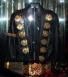 A black jacket with five round golden medals on its left and right shoulder and a gold ban on its left arm sleeve. The jacket has two belt straps on the right bottom sleeve. Underneath the jacket is a golden belt, with a round pendant in the center of it. There is a red light reflecting on the jacket and belt as well as a gold squared plat on the left side of the jacket and belt.