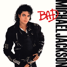 A man in his late twenties stands and looks forward. His hair is curly and black. He is wearing a black jacket that has several buckles and pants. The background is white and beside him are the words "Michael Jackson" in black capital letter, and over them, "Bad" in red.