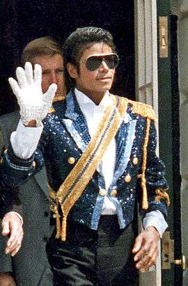 Michael Jackson wearing a sequined military jacket and dark sunglasses. He is walking while waving his right hand, which is adorned with a white glove. His left hand is bare.