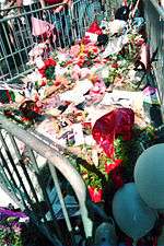 The floor on an area on the ground is covered with flowers, cards and balloons. The area is closed off with metal barricades.