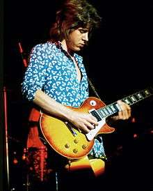 Mick Taylor playing electric guitar onstage in a blue shirt and yellow pants