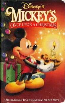 A short anthropomorphic male mouse standing at the left side of the image is holding a candle holder with a brightly burning candle, positioned on the right side of the image, with both of his hands. The mouse is wearing red shorts with white buttons and yellow shoes and is happily smiling. In the background on the left side of the picture stands a decorated Christmas tree with colorfully wrapped gifts lying under it. The video cover includes the film and company's title.