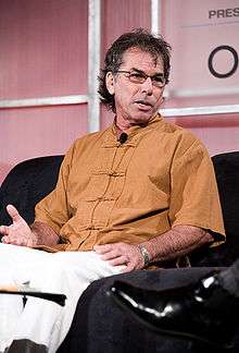 A man in an orange=colored shirt and white pants, sitting; he is wearing eyeglasses along with a microphone on his collar