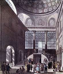 People gather in groups in a very high hall with arches, high windows, and staircases.