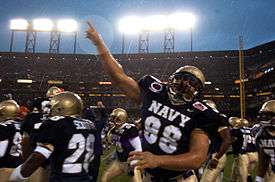 An American football player in uniform points to the sky in rainy conditions under stadium lights.