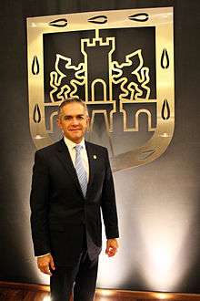 Miguel Ángel Mancera stands in front of a golden shield depicting an old Mexico City's shield. He looks directly to the camera. He wears a black suit.