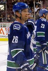 A Caucasian ice hockey player in his mid-thirties. He wears a blue jersey with white and green trim and a blue, visored helmet. He looks forward with his mouth slightly parted in a relaxed stance.