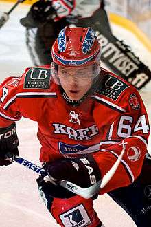An ice hockey player skating towards the camera with his ice hockey stick raised of the ice. He is wearing a red helmet and uniform.