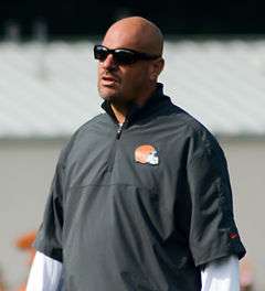 Candid waist-up photograph of Pettine wearing a grey pullover bearing a Cleveland Browns logo and dark sunglasses standing on a football field