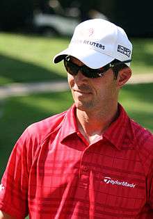 Upper body of a man in his thirties. He is wearing a red golf shirt, white baseball cap and dark sunglasses.