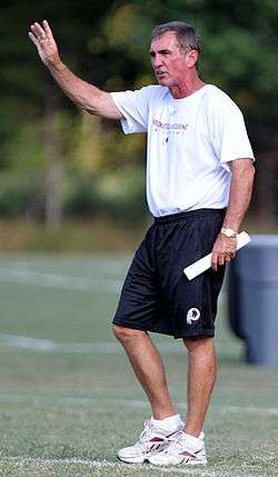 Photograph of Shanahan on a football field wearing a white Washington Redskins t-shirt and dark-colored Redskins shorts holding a rolled up sheet of paper in his left hand a raising his right hand above his head