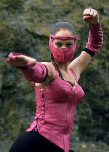 This image shows a masked woman with braided black hair, in a martial arts pose, wearing a pink-and-black outfit.