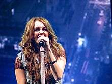 A teenage female with long, brunette hair, a fishnet shirt and a jean vest sings into a microphone. Behind her, images of a cityscape are projected.