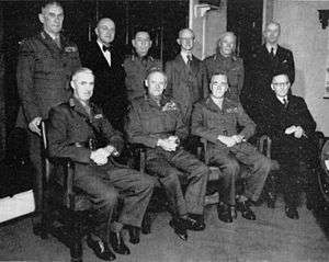 Formal portrait of a group of nine men. Four are sitting at the front and five are standing at the back. Six are wearing uniforms without headgear, while the other three are wearing civilian suits.