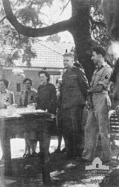 Black and white photograph of a group of three women and two men wearing military uniform. All members of the group are standing under a tree and are looking at a table.