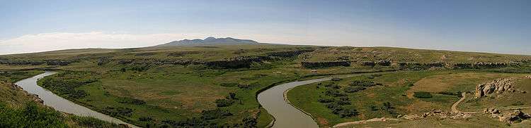 A small river winds through a rocky, grass-covered plateau. Hills rise in the distance.