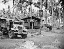 Two shirtless men in the back of a truck, driven (on the right hand side) by a third. In the background are huts on stilts with thatched rooves. The walls are only half height, leaving most of it open to the air.