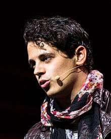A photo of Milo Yiannopoulos at the LeWeb13 Conference in Paris, France, wearing a microphone headset and a red, black, and white scarf over a brown leather jacket
