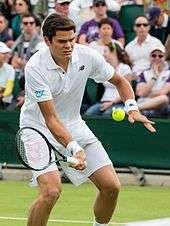 Raonic dressed in all white, bending forward slightly. His racquet is in his right hand, below the ball, about to make contact.