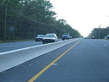 A four lane road in a wooded area with a small concrete barrier in the center. Power lines are on either side of the road.