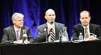 From left to right, a bespectacled middle aged man, a younger bald man holding a wireless microphone, and a balding middle aged man, all in suits, sit at a long dark table in front of dark blue curtains.