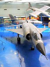 Slightly-angled front view of jet aircraft on blue floor in museum. Two aircraft are in the background, one of which is suspended near the ceiling.