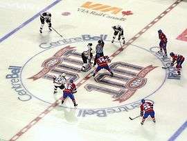 Several ice hockey players in red uniforms line up around the near half of a large circle on the ice, opposed on the other side by several in white uniforms. A player from each team stands at the centre of the circle, joined by a referee wearing a vertically striped black and white uniform.
