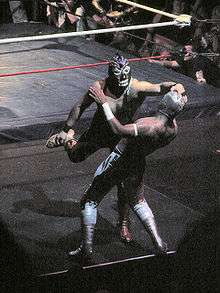 Two masked wrstlers fighting outside the ring, with one wrestling trying to pull the mask off the other.