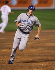 A man in a gray baseball uniform with "KANSAS CITY 35" on the chest and a blue batting helmet runs on the infield dirt of a baseball field.