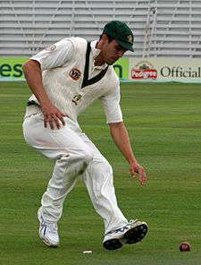 Mitchell Johnson is bending down to pick up a red cricket ball with his left hand. He is on the run.