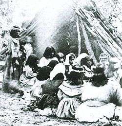 A group sits in front of a fire with a teepee made of large branches in the background.