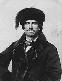 Black and white photograph of a man with a short moustache and earrings, wearing a fur lined dress jacket, bow tie and fur hat