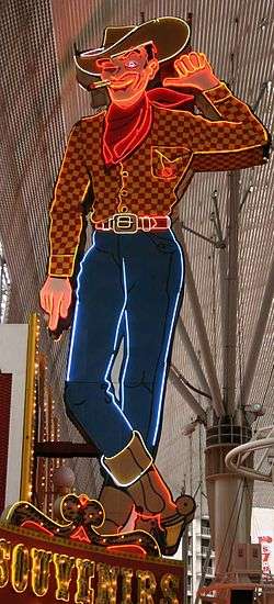 Photograph of a large painted sign in the form of a cowboy. The cowboy is winking one eye. He's lifting his left hand, and pointing that thumb towards the building to his right. A lighted cigarette dangles from the corner of his mouth. He's wearing a cowboy hat, boots, and a scarf. Glowing neon tubes highlight the outlines of the cowboy, the cigarette, his boots, and the folds of his tied scarf.