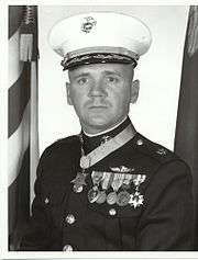 alt=A black-and-white photo of Modrzejewski from the waist up in his military dress blue uniform with hat and medals. There is an American flag and a Marine Corps flag in the background and his Medal of Honor is around his neck.