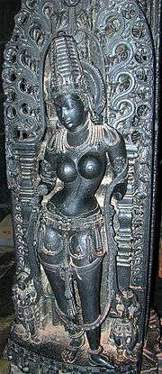 A black stone sculpture of a young topless, standing woman, wearing a conical crown with a halo and various ornaments around her waist, necklaces, bangles, bracelets, earrings, anklets as well as the sacred thread. Her arms are broken. Two small attendants stand with folded hands near her feet.