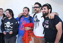 Five people. On the extreme left one person standing with a gray shirt and black jacket. Beside a person with semi-long hair, wearing a black sweatshirt with the caption "St. Pauli" holding a passport in his hands. At the center a person wearing blue shirt, holding a red jacket with black boxes. At his side a person with dark glasses, beard, wearing a white shirt with a black legend and brown pants with red belt. To the far right a person standing with dark glasses on his head, bearded and wearing a black colored shirt.