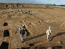 A photograph of a wide flat area of dirt, with a handful of men scattered across it engaged in some sort of work. In the near ground one man is standing looking at the camera. All the men cast long shadows across the picture.