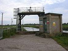 A metal gantry between the road in the foreground and a river. To the right is a breeze block building with warning signs on it.